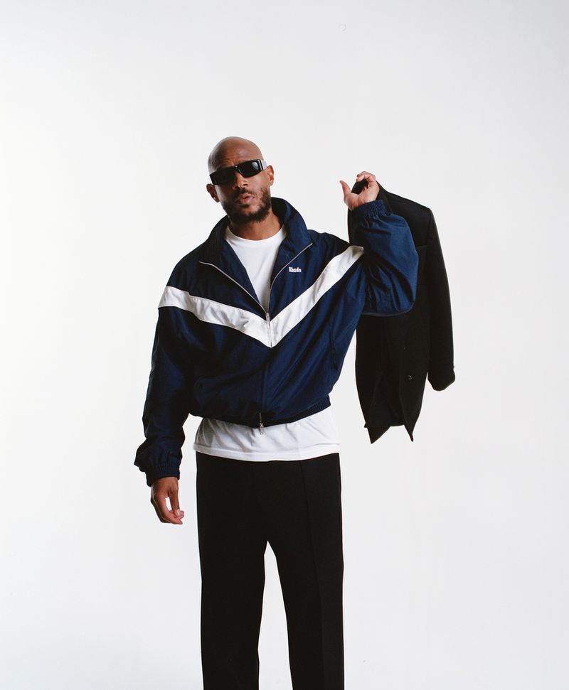 Bald man in stylish track jacket and sunglasses holds a black suit jacket in a bright studio environment. Fashion and lifestyle.