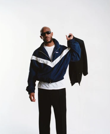 Bald man in stylish track jacket and sunglasses holds a black suit jacket in a bright studio environment. Fashion and lifestyle.