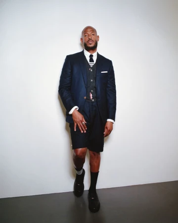 A man in a navy suit, vest, tie, and black shorts stands against a white wall in a fashion studio. Full shot, eye-level angle.