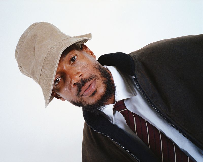 Portrait of an adult male wearing a bucket hat, white dress shirt, tie, and jacket, posing in a photo studio with a plain backdrop.
