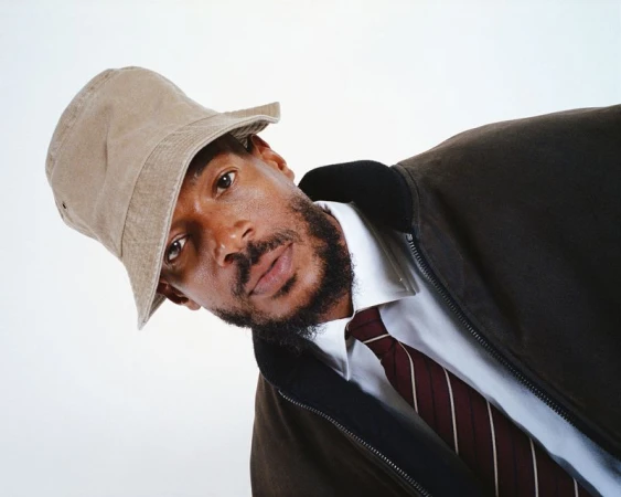 Portrait of an adult male wearing a bucket hat, white dress shirt, tie, and jacket, posing in a photo studio with a plain backdrop.