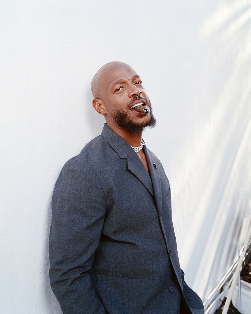 Bald man in gray suit with cigar poses against white wall outdoors, wearing necklace and looking at camera with slight smile.
