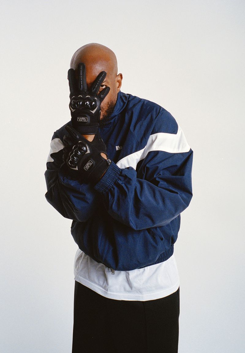 A bald man in a blue windbreaker jacket, black pants and black gloves is in a medium shot in a brightly lit photo studio.