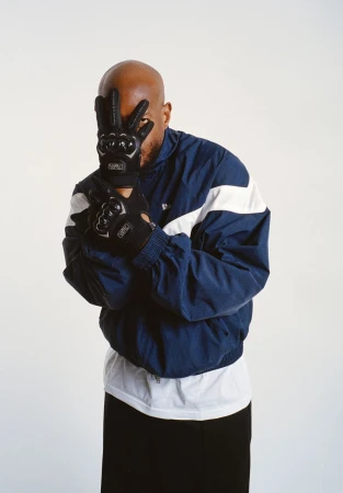A bald man in a blue windbreaker jacket, black pants and black gloves is in a medium shot in a brightly lit photo studio.