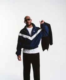 Swizz Beatz wearing sunglasses, a blue track jacket, and black trousers while holding a coat over his shoulder on a white backdrop.