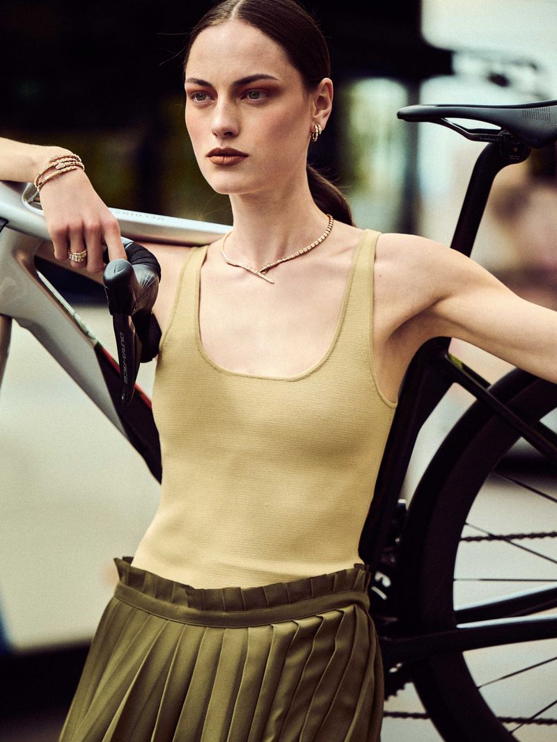 Woman in a yellow top and green skirt poses with a grey racing bicycle on city streets, wearing jewelry and neutral makeup.