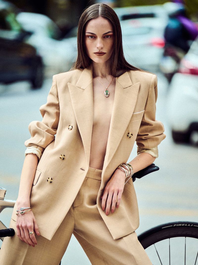 Fashion editorial shot of a female model in a beige suit, posing with a bicycle on a city street with cars blurred in the background.