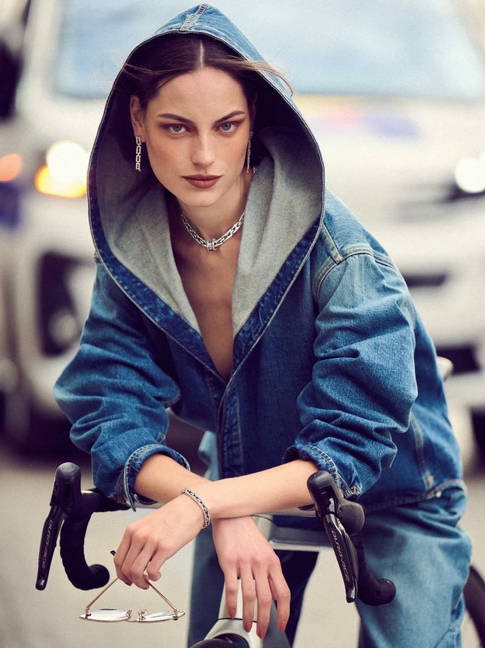 Model in denim hooded jacket leaning on bicycle handlebars with silver jewellery against blurred street