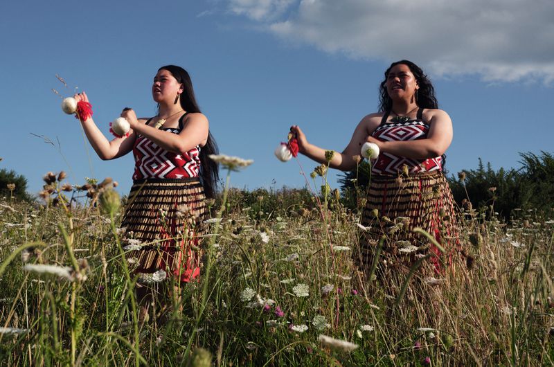 Two adult women in traditional Maori clothing perform with poi in an outdoor field of wildflowers against a partly cloudy blue sky.