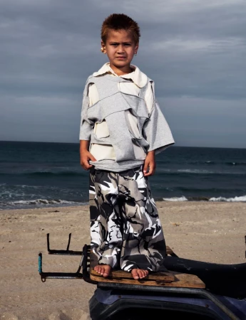 A young child wearing a patterned top and pants stands barefoot on a beach next to the ocean under a cloudy sky, looking at the camera.
