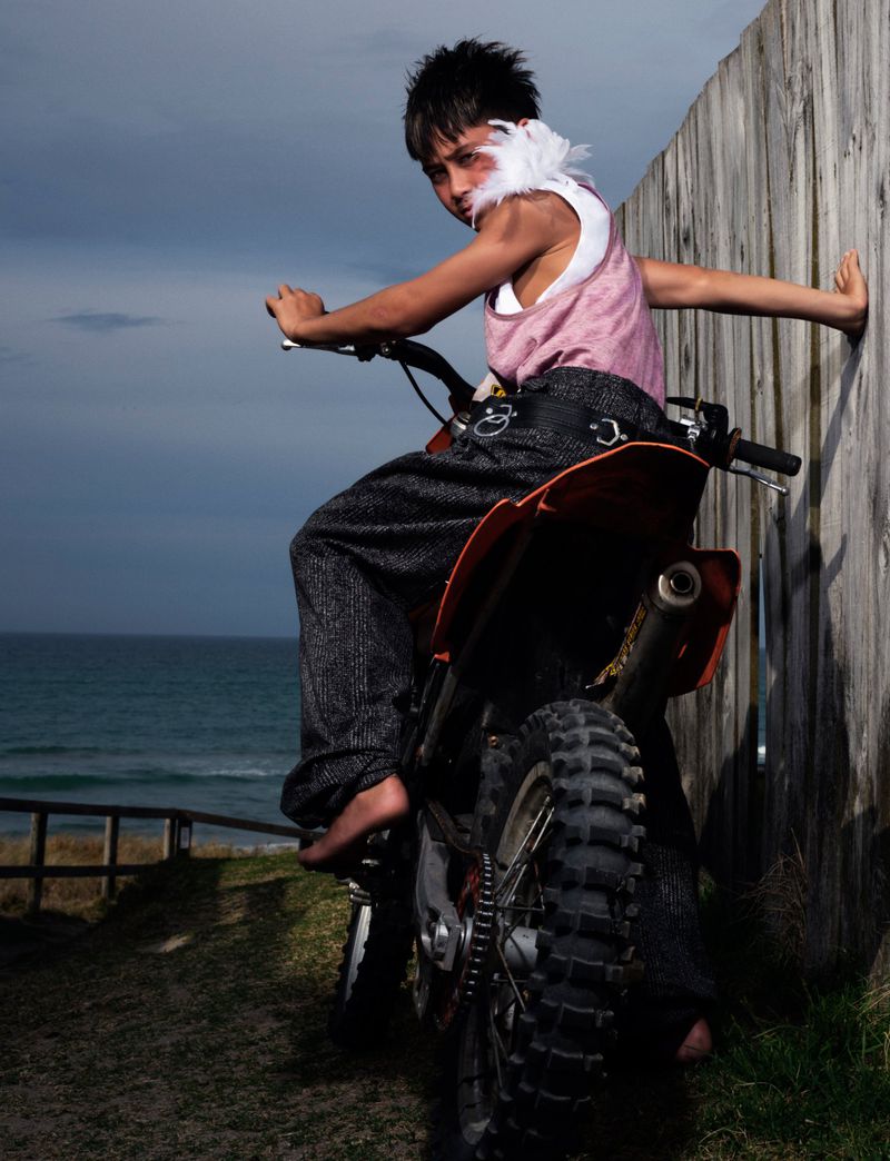 Young barefoot male child with feathers poses on orange dirt bike by a wooden fence near the ocean in an outdoor setting.