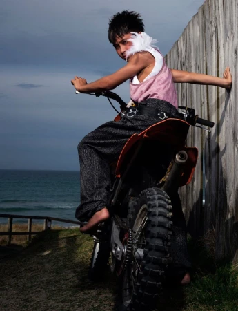 Young barefoot male child with feathers poses on orange dirt bike by a wooden fence near the ocean in an outdoor setting.