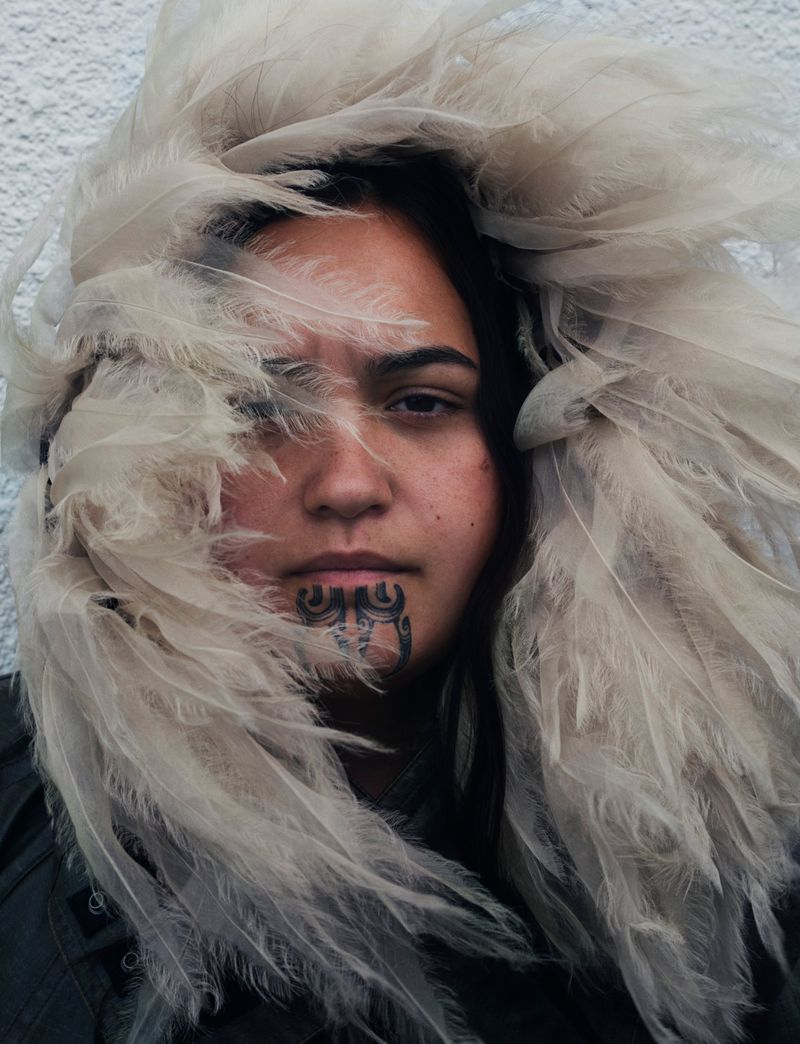 Portrait of a woman with a chin tattoo surrounded by white feathers in an indoor setting with a neutral background.