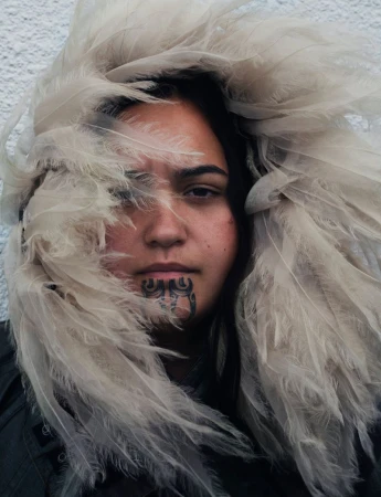 Portrait of a woman with a chin tattoo surrounded by white feathers in an indoor setting with a neutral background.