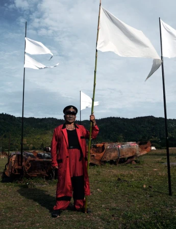 An older adult stands in a field holding a flag, wearing a red coat and hat, with a landscape backdrop, burned vehicles, and white flags.