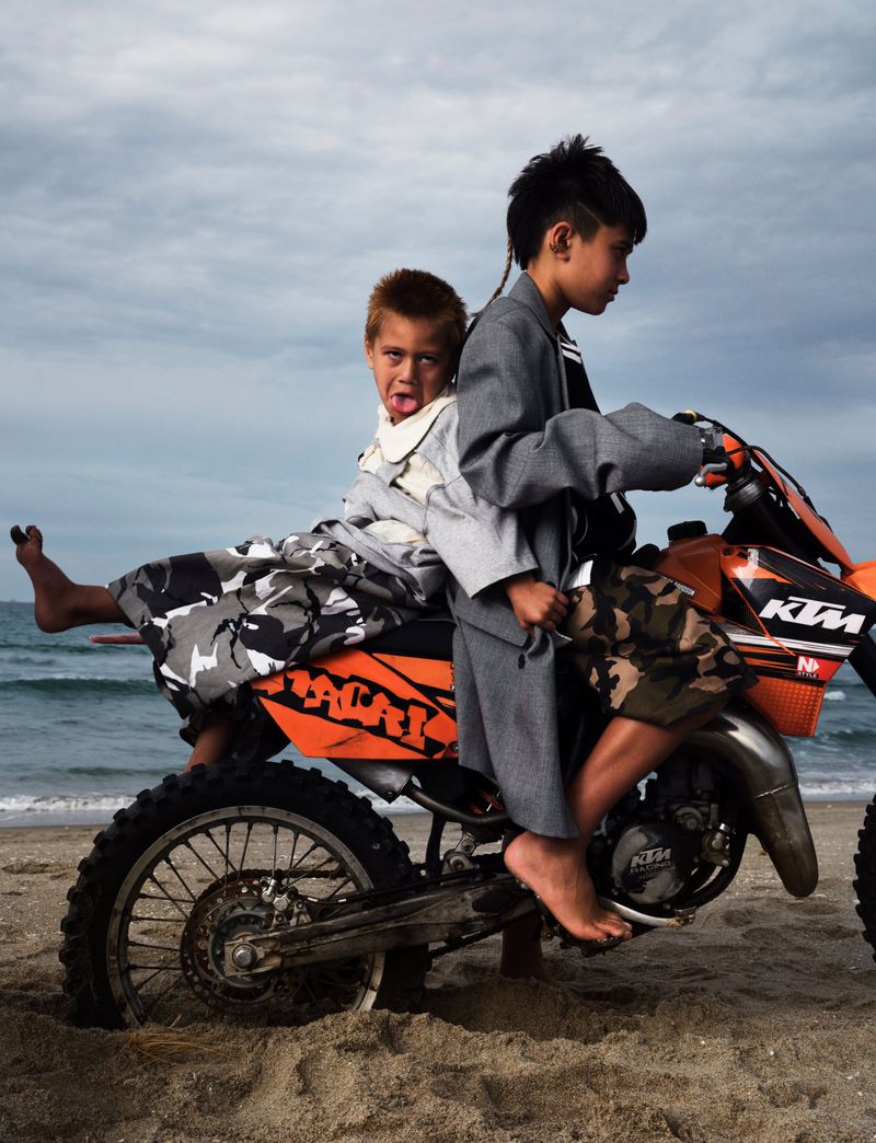 Two children ride an orange and black dirt bike on a beach, with a cloudy sky and ocean backdrop.