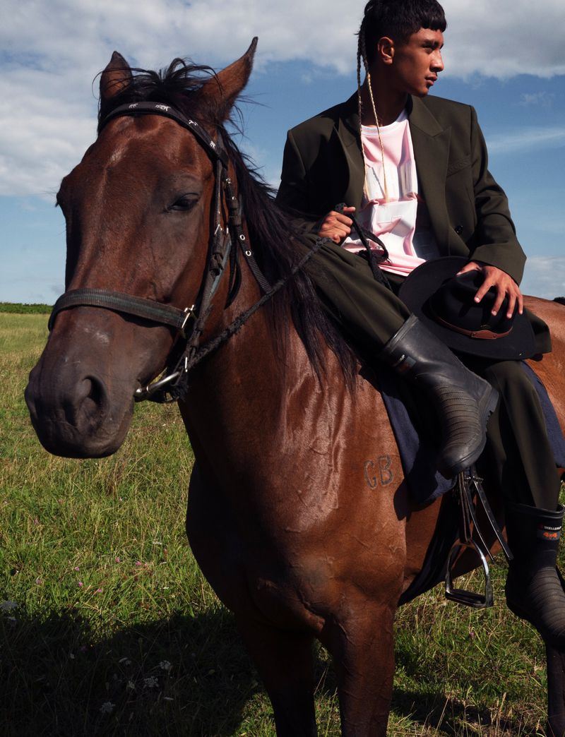 Model sits astride a brown horse outdoors in a field wearing a dark suit and black riding boots, holding a black hat with a blue sky.