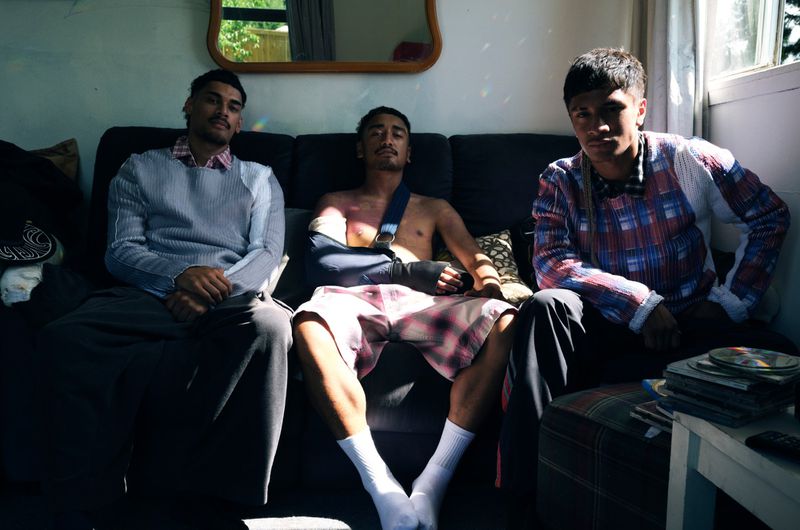 Three young adult men sit on a dark couch in front of a window. One man has an arm in a sling. Sunlight casts shadows across the scene.