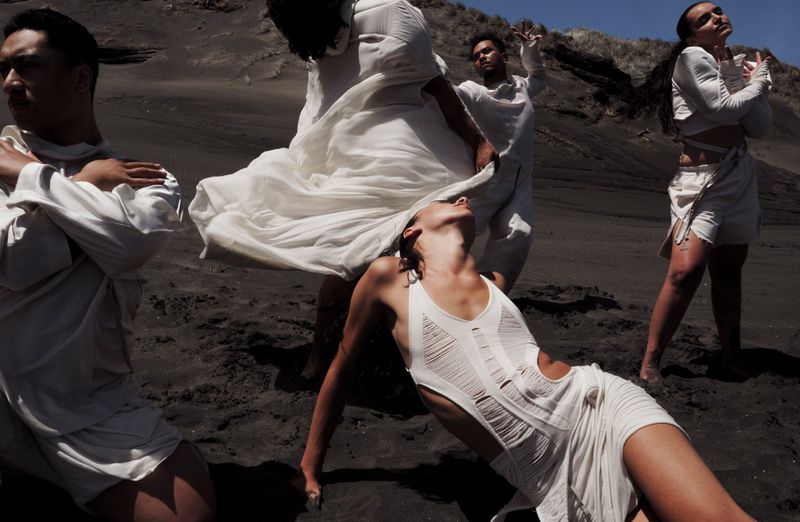 A group of people dressed in white clothing posing on a dark sandy beach with a hilly background on a sunny day, medium shot.