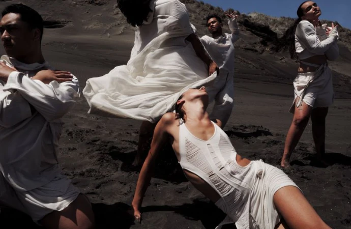 A group of people dressed in white clothing posing on a dark sandy beach with a hilly background on a sunny day, medium shot.