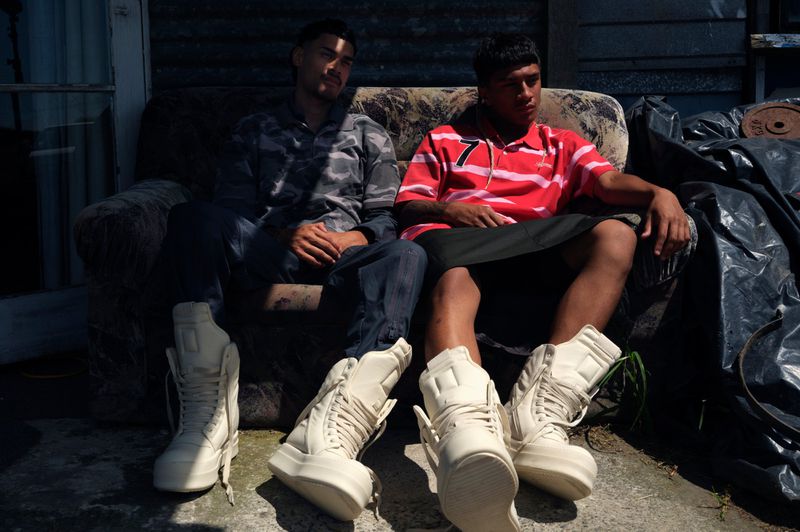 Two men in casual clothes sitting on a weathered sofa outdoors, both wearing platform sneakers. The scene is set in a rustic environment.