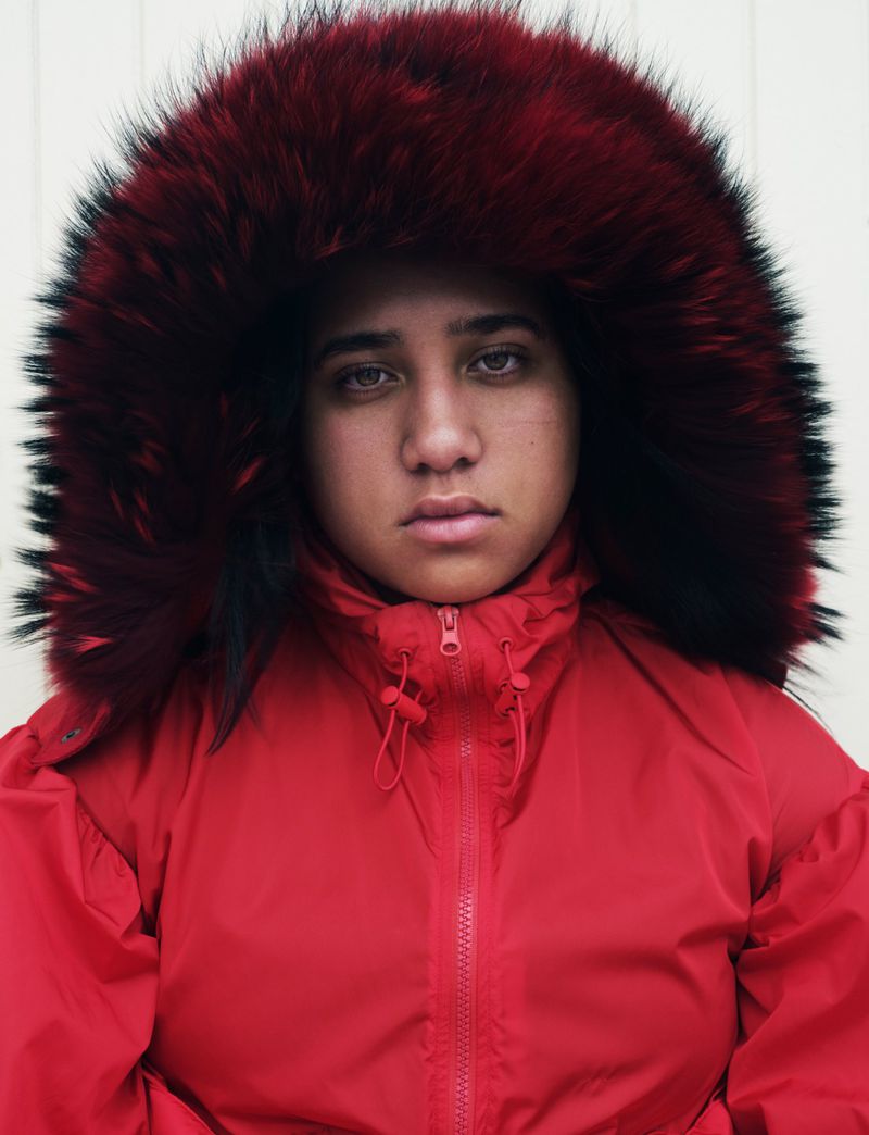 Portrait of a young person with fair skin, wearing a red puffer coat with a fur-lined hood, gazing directly at the camera.