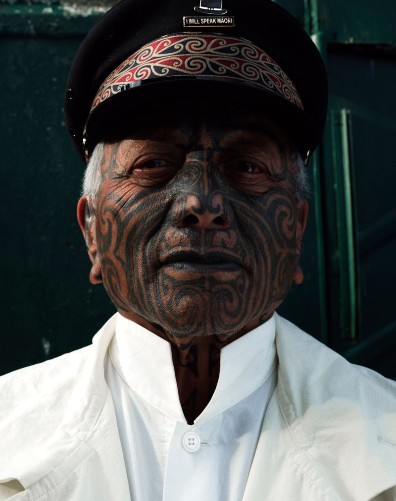 Close-up of a senior Maori man with traditional facial tattoos, wearing a cap and white jacket, against a solid dark-green background.