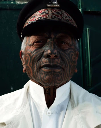 Close-up of a senior Maori man with traditional facial tattoos, wearing a cap and white jacket, against a solid dark-green background.