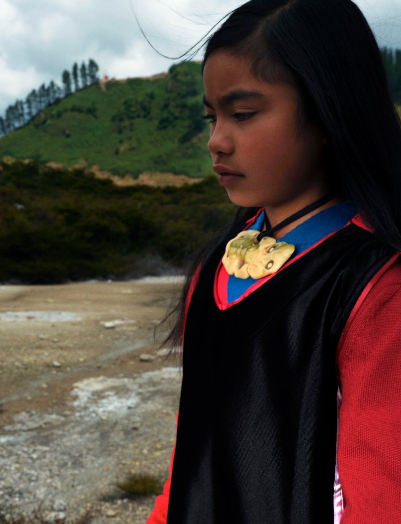 Young Maori girl in traditional clothing stands outdoors against a blurred background of geothermal terrain and a distant mountain.