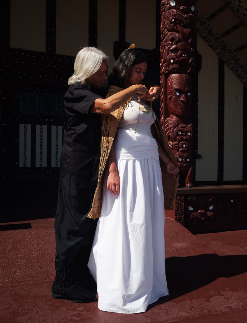 Two women standing outdoors, one assisting the other with a woven shawl and necklace, next to a carved wooden post and building.