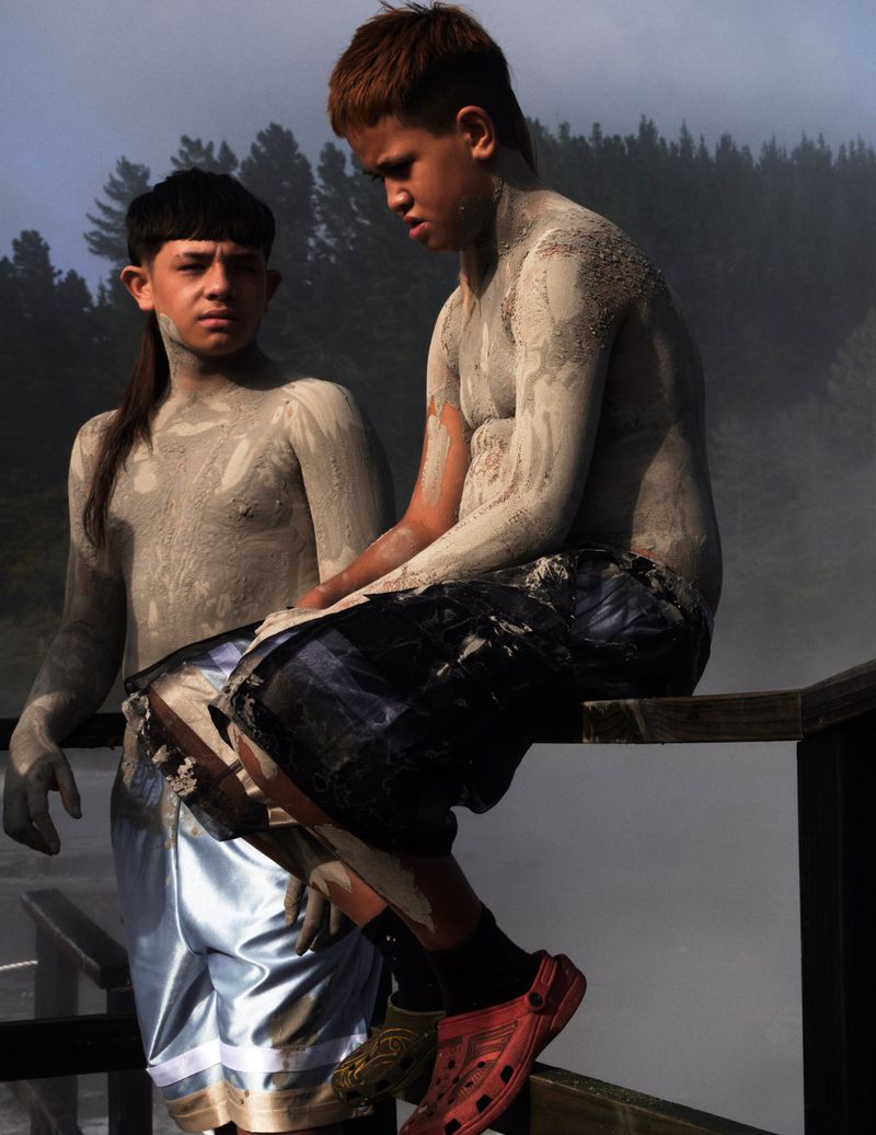 Two boys covered in mud sit on a wooden fence in a hazy outdoor setting with visible trees. They wear shorts and casual shoes.