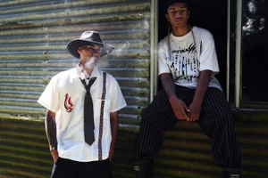 Two young men posing outdoors against a corrugated metal wall, one wearing a hat and smoking while the other sits in a doorway