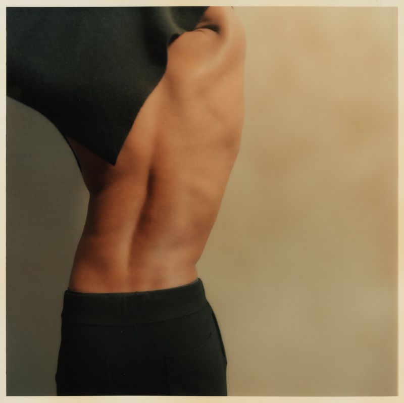 Torso and back of a slender adult female with dark hair covered by black cloth, wearing black bottoms, against a light beige background.
