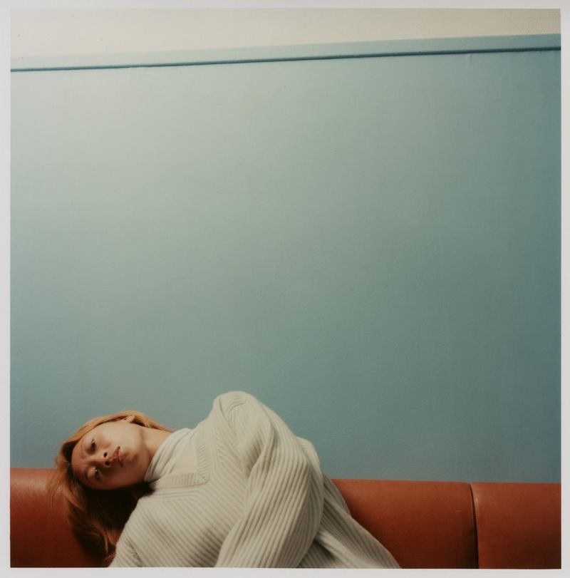 Adult female laying on an orange couch indoors with a serene expression in front of a light blue wall and white sweater.