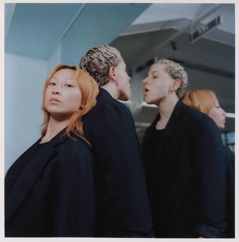Two adult women pose in dark jackets in front of a mirror, creating a mirrored effect with four figures in a minimalist indoor setting.
