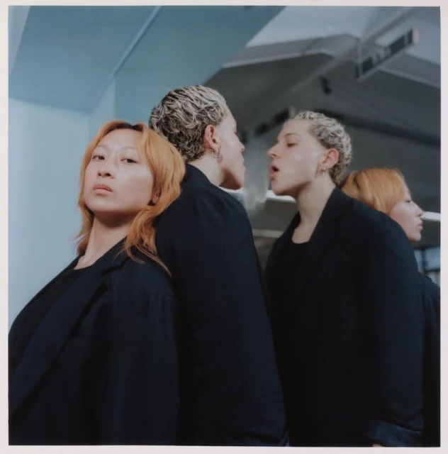 Two adult women pose in dark jackets in front of a mirror, creating a mirrored effect with four figures in a minimalist indoor setting.