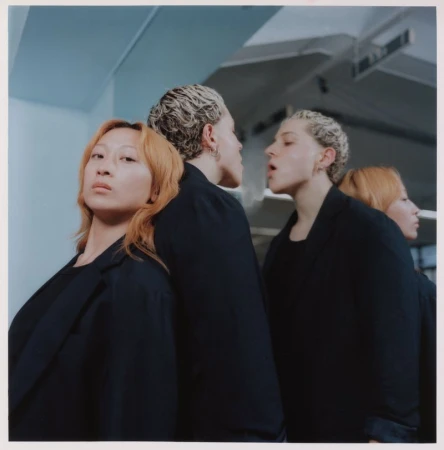 Two adult women pose in dark jackets in front of a mirror, creating a mirrored effect with four figures in a minimalist indoor setting.
