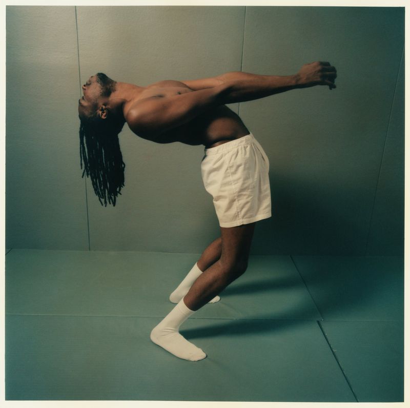 Athletic African-American man in a backbend pose wearing white shorts and socks against a neutral studio backdrop, arms extended.