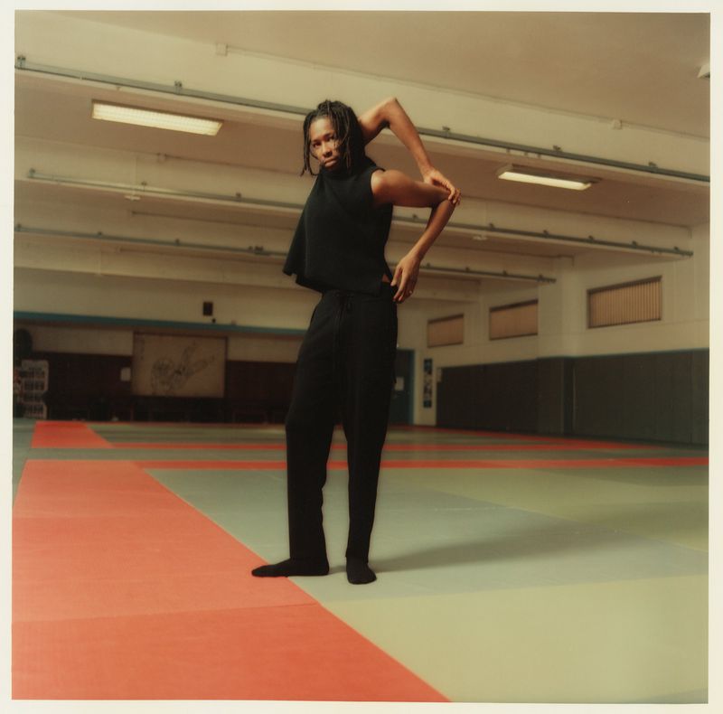 An adult in a black top and pants in a dance studio stretches their arms, standing on a red and green floor mat.