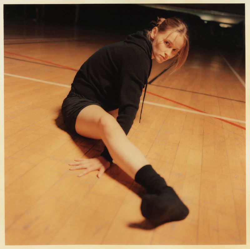 Female dancer in black hoodie and shorts stretches on wood gym floor with white lines, looking directly at camera with focused expression.