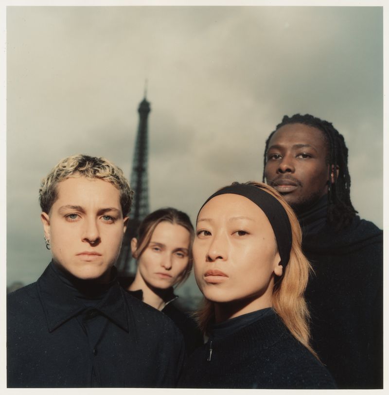 Four people in black attire pose in front of the Eiffel Tower in Paris, France, with a cloudy sky providing a subdued backdrop.