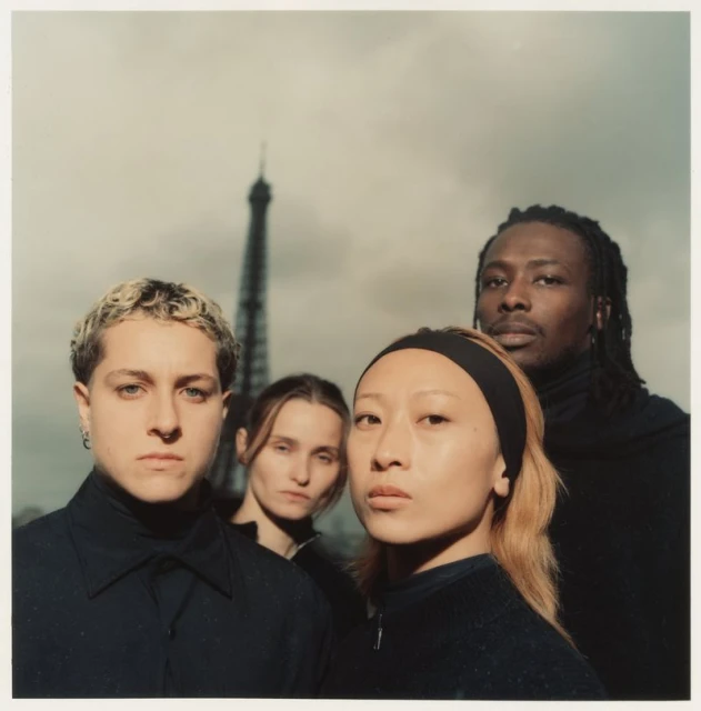 Four people in black attire pose in front of the Eiffel Tower in Paris, France, with a cloudy sky providing a subdued backdrop.