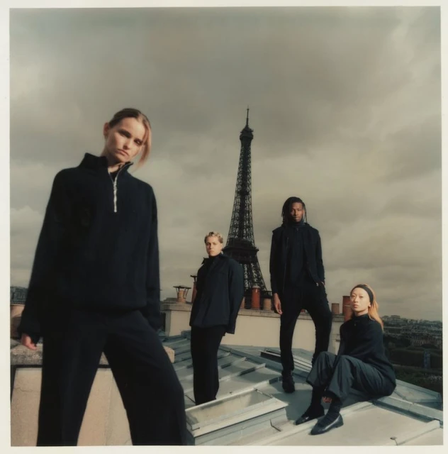 Four fashion models in black clothing pose on a rooftop with the Eiffel Tower in the background on an overcast day in Paris.