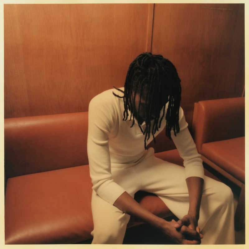Adult with dark hair wearing a cream-colored outfit sitting on an orange bench against a matching wall indoors with natural lighting.