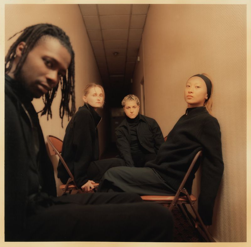 Group of four diverse young adults seated in chairs in dimly lit corridor. Three women and one man, all in dark clothing, posing indoors.