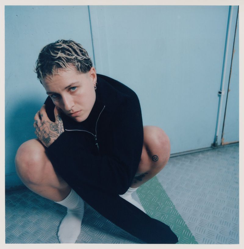 Eye-level shot of a young adult with short bleached hair crouching indoors with a light blue wall and a textured metal floor.