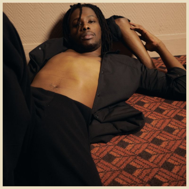 An African American man with dreadlocks poses in a relaxed manner, lying shirtless with an unbuttoned black shirt, on patterned flooring.