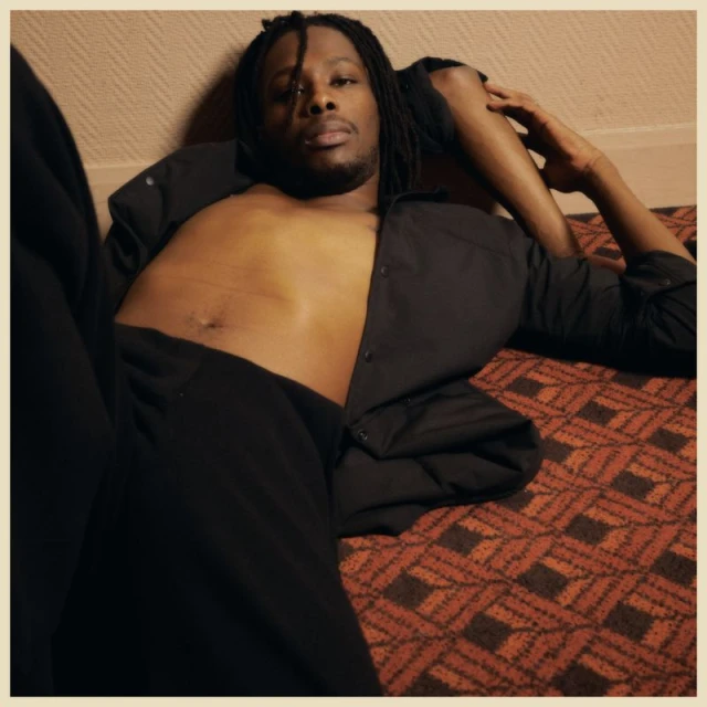 An African American man with dreadlocks poses in a relaxed manner, lying shirtless with an unbuttoned black shirt, on patterned flooring.