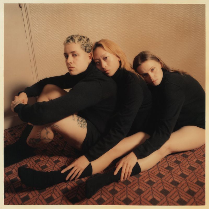 Three diverse young adults in black turtlenecks pose together on a patterned rug, sitting close and looking thoughtfully at the viewer.