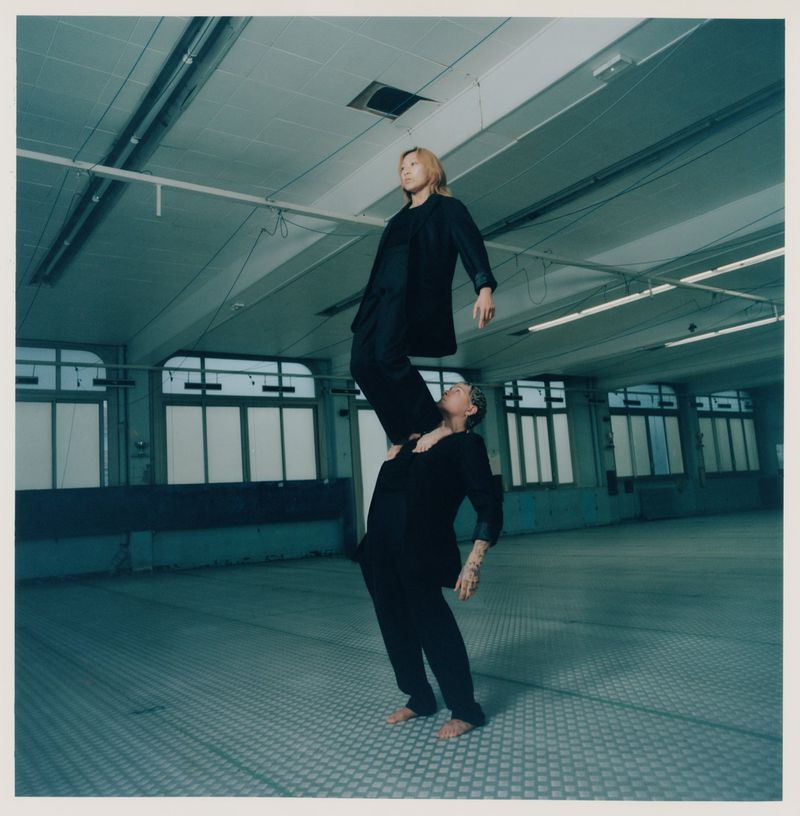 A man balances a woman on his shoulders in a minimalist industrial setting, both in black suits, in a square shot.